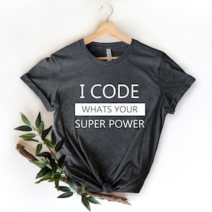 Peut inclure: T-shirt gris foncé avec le texte blanc "I CODE WHATS YOUR SUPER POWER". Le t-shirt est suspendu à un cintre en bois. Des feuilles vertes et une branche sont placées en bas à gauche du t-shirt.