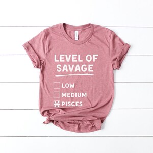 Op de afbeelding: Een stoffig roze t-shirt met de witte tekst "LEVEL OF SAVAGE", met selectievakjes voor "LOW" en "MEDIUM", en een aangevinkt vakje voor "PISCES". Het t-shirt is aan de onderkant geknoopt.
