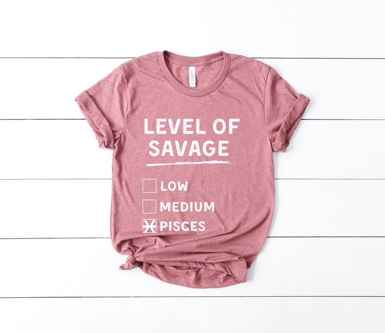 Op de afbeelding: Een stoffig roze t-shirt met de witte tekst "LEVEL OF SAVAGE", met selectievakjes voor "LOW" en "MEDIUM", en een aangevinkt vakje voor "PISCES". Het t-shirt is aan de onderkant geknoopt.