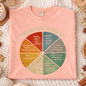 Puede incluir: Camiseta melocot&oacute;n con un dise&ntilde;o de diagrama circular y la frase "YOU MAY ASK YOURSELF". El diagrama incluye preguntas como "&iquest;C&oacute;mo llegu&eacute; aqu&iacute;?" y "&iquest;D&oacute;nde est&aacute; ese coche grande?"