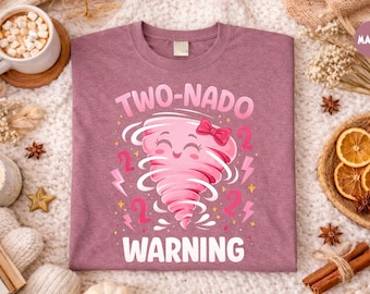 May include: Mauve-colored t-shirt with a graphic of a pink tornado with a bow, the number 2, and lightning bolts. The text reads "TWO-NADO WARNING". The shirt is on a white, textured surface.