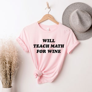 Può includere: Una maglietta rosa chiaro con la scritta "WILL TEACH MATH FOR WINE" in grassetto nero. La maglietta è appesa a un gancio di legno contro un muro bianco, con un cappello grigio sullo sfondo.