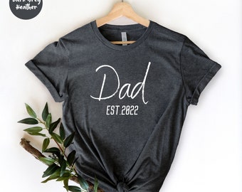 Puede incluir: Camiseta gris oscuro jaspeado con la palabra "Dad" en escritura blanca y "EST.2022" en una fuente sencilla. La camiseta está colgada de una percha de madera. Una ramita de hojas verdes y una rama están en la esquina inferior izquierda.