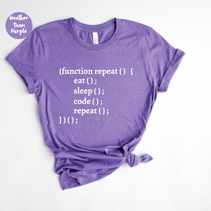 May include: A heather purple t-shirt with white text that reads "(function repeat () { eat (); sleep (); code (); repeat (); })();". The shirt has a crew neck and short sleeves. The text "Heather Team Purple" is in a circle in the upper left corner.