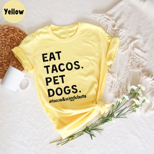 Op de afbeelding: Geel T-shirt met zwarte tekst die "EAT TACOS. PET DOGS. #tacos&wigglebutts" zegt