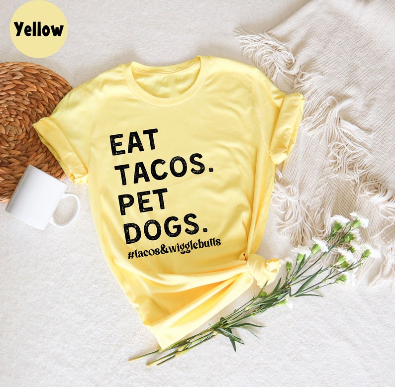 Op de afbeelding: Geel T-shirt met zwarte tekst die "EAT TACOS. PET DOGS. #tacos&wigglebutts" zegt