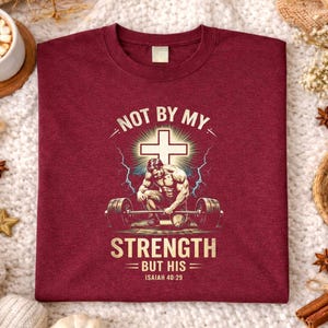 K&ouml;nnte beinhalten: Burgunderfarbenes T-Shirt mit dem Text "NOT BY MY STRENGTH BUT HIS" und einer Grafik eines Gewichthebers und eines Kreuzes. Das Shirt enth&auml;lt auch den Bibelvers Jesaja 40:29.