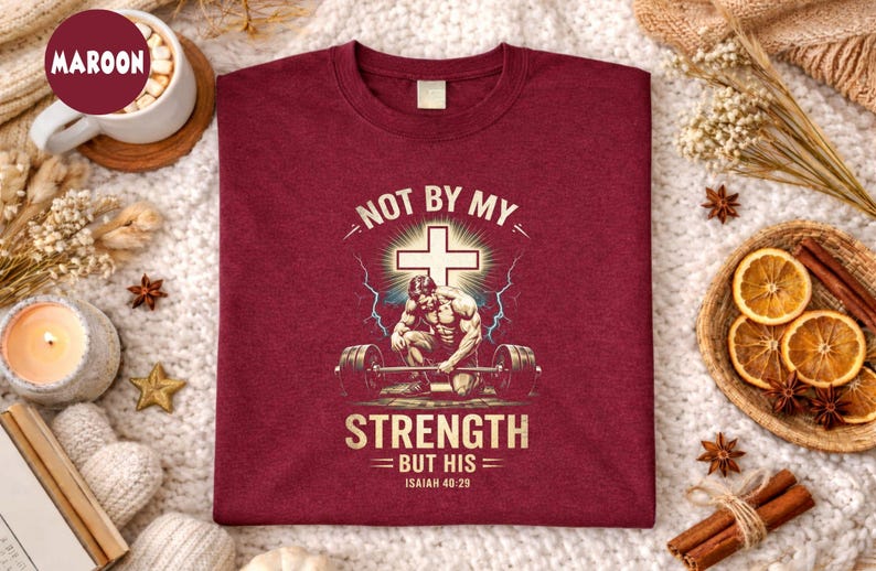 K&ouml;nnte beinhalten: Burgunderfarbenes T-Shirt mit dem Text "NOT BY MY STRENGTH BUT HIS" und einer Grafik eines Gewichthebers und eines Kreuzes. Das Shirt enth&auml;lt auch den Bibelvers Jesaja 40:29.
