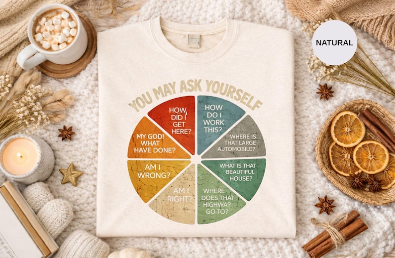 Puede incluir: Sudadera color crema con la frase "YOU MAY ASK YOURSELF" y un dise&ntilde;o de diagrama circular. El gr&aacute;fico incluye preguntas como "&iquest;C&oacute;mo llegu&eacute; aqu&iacute;?" y "&iquest;D&oacute;nde est&aacute; ese coche grande?" La palabra "NATURAL" est&aacute; en un c&iacute;rculo.