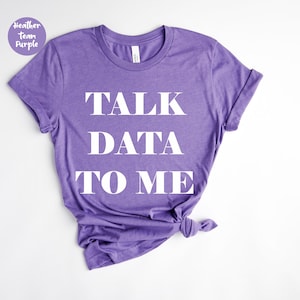 以下が含まれることがあります： 「TALK DATA TO ME」の白文字がプリントされた紫色のTシャツ。裾は結ばれています。左上に「Heather Team Purple」と書かれた小さな丸いラベルがあります。