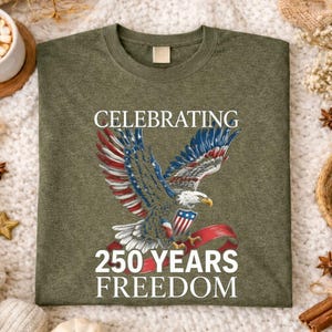 May include: Olive green t-shirt featuring a detailed graphic of an eagle with red, white, and blue wings, holding a banner. The text reads "CELEBRATING 250 YEARS FREEDOM". The shirt is displayed on a textured surface.