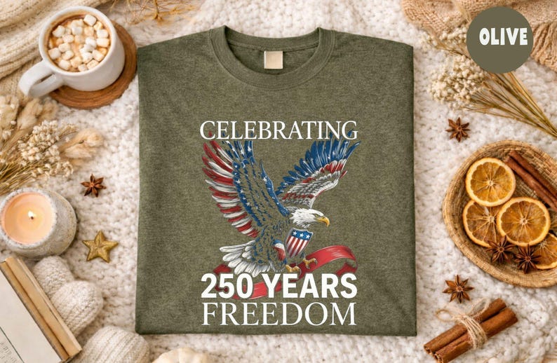May include: Olive green t-shirt featuring a detailed graphic of an eagle with red, white, and blue wings, holding a banner. The text reads "CELEBRATING 250 YEARS FREEDOM". The shirt is displayed on a textured surface.