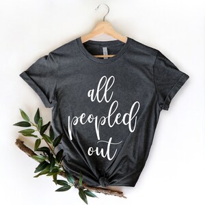 May include: Dark gray t-shirt with the white text "all peopled out." The shirt is hanging on a wooden hanger. A sprig of greenery is placed on the lower left side of the shirt.