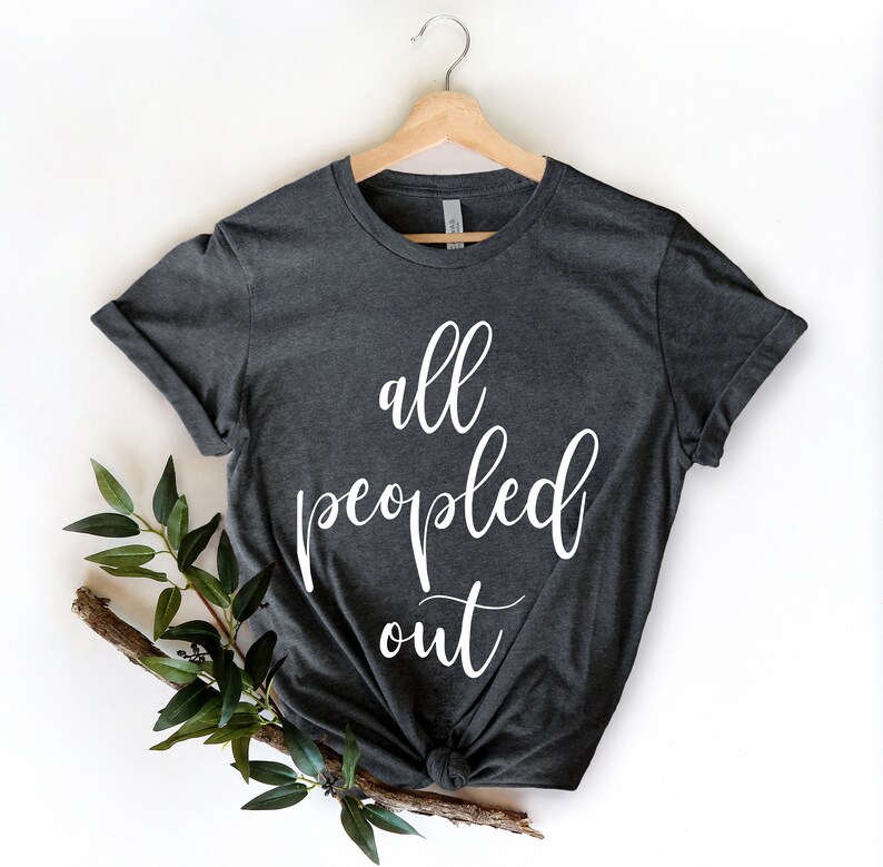 May include: Dark gray t-shirt with the white text "all peopled out." The shirt is hanging on a wooden hanger. A sprig of greenery is placed on the lower left side of the shirt.