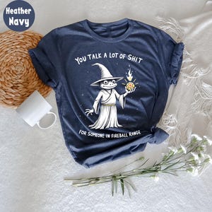 May include: Heather navy blue t-shirt featuring a frog wizard illustration holding a fireball. The text on the shirt says "YOU TALK A LOT OF SHIT FOR SOMEONE IN FIREBALL RANGE."