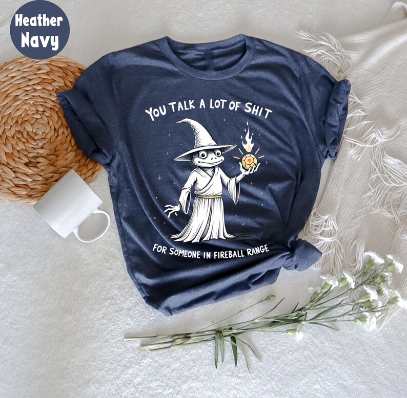 May include: Heather navy blue t-shirt featuring a frog wizard illustration holding a fireball. The text on the shirt says "YOU TALK A LOT OF SHIT FOR SOMEONE IN FIREBALL RANGE."