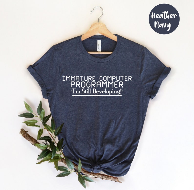 May include: Heather navy t-shirt with the text "IMMATURE COMPUTER PROGRAMMER - I'm Still Developing!" in white. The shirt is hanging on a wooden hanger with a sprig of greenery.