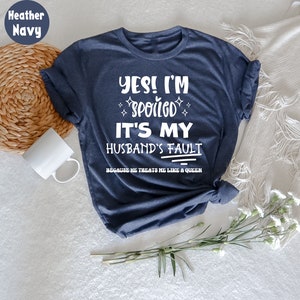 May include: A dark blue t-shirt with white text that reads "YES! I'M SPOILED IT'S MY HUSBAND'S FAULT BECAUSE HE TREATS ME LIKE A QUEEN".