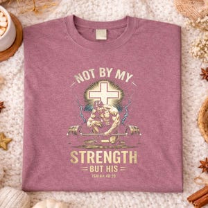 K&ouml;nnte beinhalten: Mauvefarbenes T-Shirt mit einem Grafikdesign, das ein Kreuz, einen Gewichtheber und den Text "NOT BY MY STRENGTH BUT HIS Isaiah 40:29" zeigt. Das Design ist in Gold und Wei&szlig; gehalten. Das Shirt wird auf einer strukturierten Oberfl&auml;che pr&auml;sentiert.