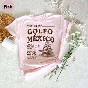 Op de afbeelding: Roze T-shirt met een bruin grafisch ontwerp met de tekst "The Name Golfo de Mexico", een vintage scheepsafbeelding en tekst over de Golf van Mexico die in 1550 op een wereldkaart verscheen.