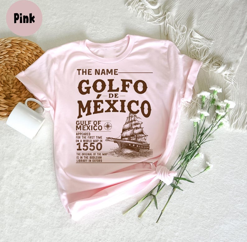 Op de afbeelding: Roze T-shirt met een bruin grafisch ontwerp met de tekst "The Name Golfo de Mexico", een vintage scheepsafbeelding en tekst over de Golf van Mexico die in 1550 op een wereldkaart verscheen.