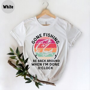 May include: White t-shirt with a colorful graphic of a fish jumping out of water, set against a striped sunset design. The text "GONE FISHING" arches above the fish, with "BE BACK AROUND WHEN I'M DONE O'CLOCK" below. The shirt is displayed on a wooden hanger.