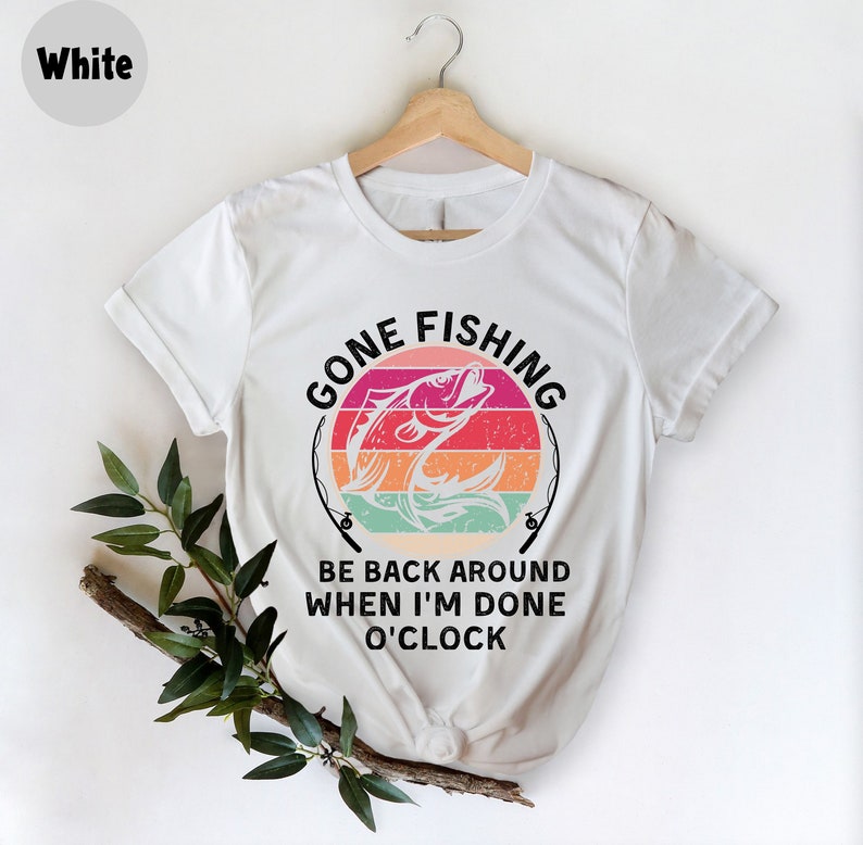 May include: White t-shirt with a colorful graphic of a fish jumping out of water, set against a striped sunset design. The text "GONE FISHING" arches above the fish, with "BE BACK AROUND WHEN I'M DONE O'CLOCK" below. The shirt is displayed on a wooden hanger.