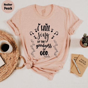 K&ouml;nnte beinhalten: Hellrosa T-Shirt mit dem Text "I WILL SING OF THE GOODNESS OF GOD" und Noten. Das Shirt enth&auml;lt auch die Bibelstelle Psalm 89,1. Eine Kaffeetasse, eine Pflanze und ein Notizbuch befinden sich im Hintergrund.