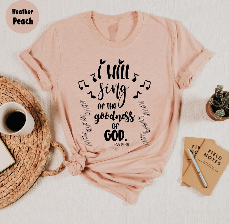 K&ouml;nnte beinhalten: Hellrosa T-Shirt mit dem Text "I WILL SING OF THE GOODNESS OF GOD" und Noten. Das Shirt enth&auml;lt auch die Bibelstelle Psalm 89,1. Eine Kaffeetasse, eine Pflanze und ein Notizbuch befinden sich im Hintergrund.