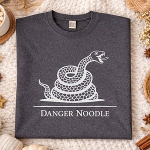 Peut inclure: Sweat-shirt gris chin&eacute; fonc&eacute; avec un motif blanc d'un serpent enroul&eacute;, la langue sortie. Le texte "DANGER NOODLE" est en dessous. Le sweat-shirt est pr&eacute;sent&eacute; sur une surface blanche textur&eacute;e avec des &eacute;l&eacute;ments d&eacute;coratifs.