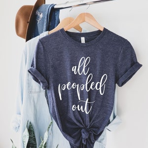 May include: A navy blue t-shirt with the words "all peopled out" in white script. The shirt is knotted at the bottom and hangs on a wooden hanger. A light blue button-down shirt and a brown hat are also visible.