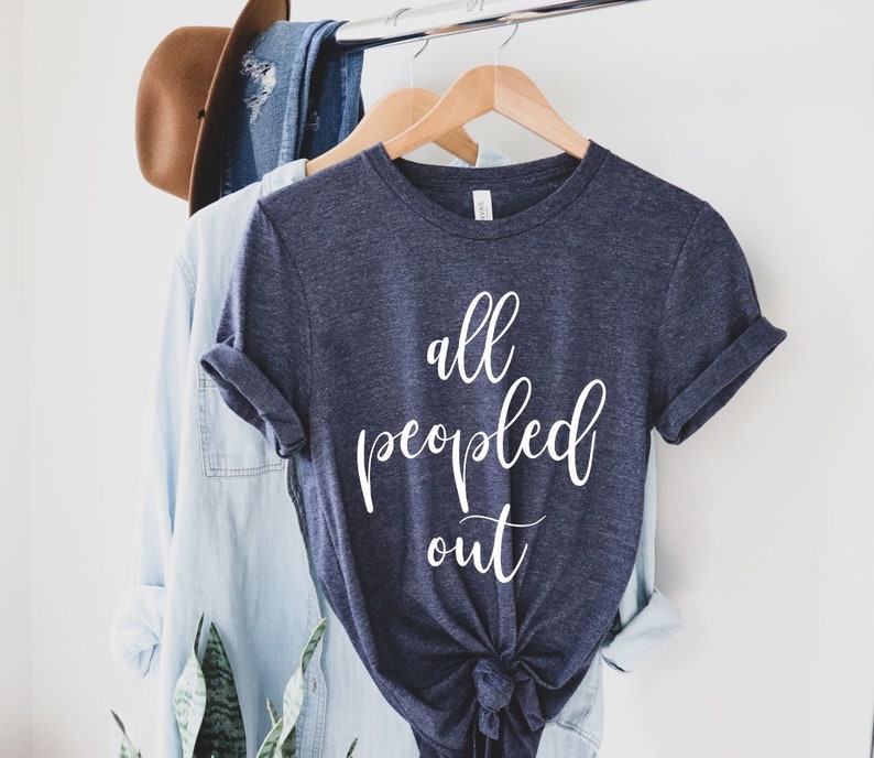 May include: A navy blue t-shirt with the words "all peopled out" in white script. The shirt is knotted at the bottom and hangs on a wooden hanger. A light blue button-down shirt and a brown hat are also visible.