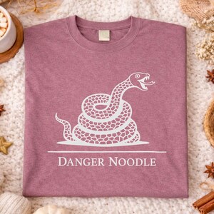 Peut inclure: T-shirt mauve avec un motif blanc d'un serpent enroul&eacute;, la langue sortie. Le texte "DANGER NOODLE" est en dessous. Le t-shirt est pr&eacute;sent&eacute; sur une surface textur&eacute;e avec des &eacute;l&eacute;ments d&eacute;coratifs.