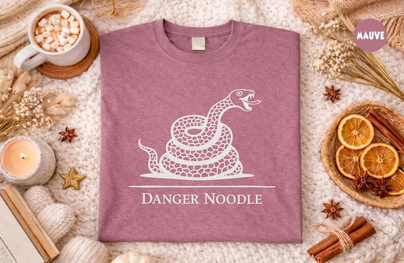 Peut inclure: T-shirt mauve avec un motif blanc d'un serpent enroul&eacute;, la langue sortie. Le texte "DANGER NOODLE" est en dessous. Le t-shirt est pr&eacute;sent&eacute; sur une surface textur&eacute;e avec des &eacute;l&eacute;ments d&eacute;coratifs.