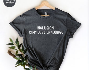 Pode incluir: T-shirt cinza escuro mesclado com o texto branco "INCLUSION IS MY LOVE LANGUAGE". A camiseta é exibida em um cabide de madeira contra um fundo branco. A vegetação é colocada no canto inferior esquerdo.