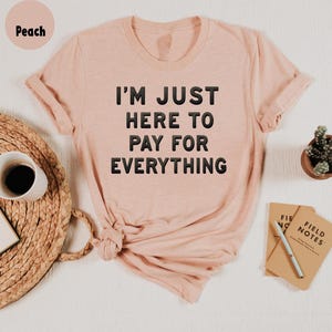K&ouml;nnte beinhalten: Ein T-Shirt in Pfirsichfarbe mit dem Aufdruck "I'm just here to pay for everything" in Schwarz.