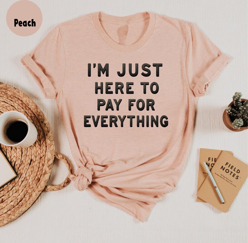 K&ouml;nnte beinhalten: Ein T-Shirt in Pfirsichfarbe mit dem Aufdruck "I'm just here to pay for everything" in Schwarz.