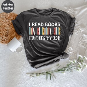May include: Dark grey heather t-shirt with the text "I READ BOOKS LIKE IT'S MY JOB" in white. The design features a row of colourful books. A white mug and a woven basket are visible in the background.
