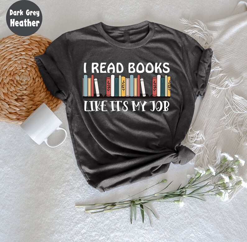 May include: Dark grey heather t-shirt with the text "I READ BOOKS LIKE IT'S MY JOB" in white. The design features a row of colourful books. A white mug and a woven basket are visible in the background.