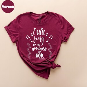 K&ouml;nnte beinhalten: Burgunderfarbenes T-Shirt mit dem wei&szlig;en Text &bdquo;I will sing of the goodness of God&ldquo; und Noten. Der Bibelvers Psalm 89,1 ist darunter aufgedruckt. Das T-Shirt ist gefaltet und am unteren Ende geknotet.