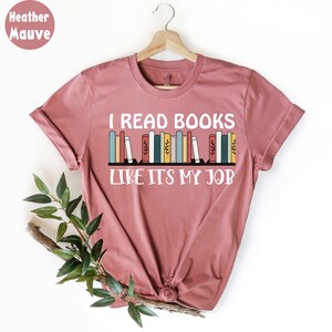 May include: Mauve t-shirt with the text "I READ BOOKS LIKE ITS MY JOB" in white. A graphic of colourful books is above the text. The shirt is displayed on a wooden hanger.