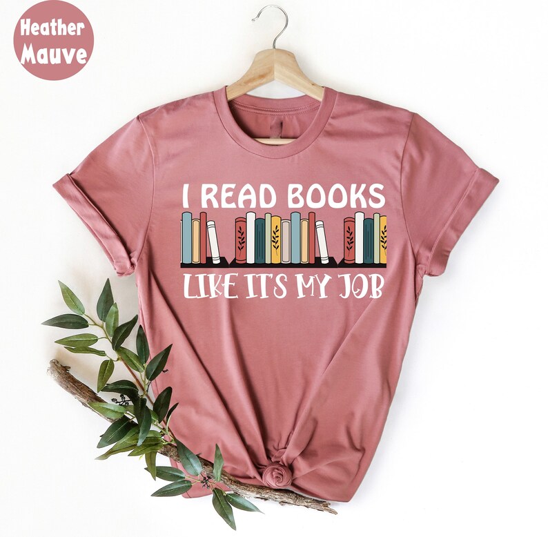 May include: Mauve t-shirt with the text "I READ BOOKS LIKE ITS MY JOB" in white. A graphic of colourful books is above the text. The shirt is displayed on a wooden hanger.