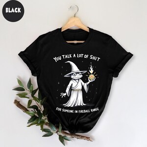 May include: Black t-shirt with a cartoon wizard frog design and the text "YOU TALK A LOT OF SHIT FOR SOMEONE IN FIREBALL RANGE." The frog is dressed in a white robe and hat, holding a glowing orb.