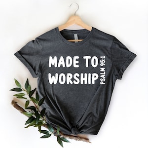 May include: Dark grey t-shirt with the words "MADE TO WORSHIP" in white block letters. The text "PSALM 95:1" is printed vertically on the right side. The shirt is hanging on a wooden hanger.