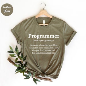 Puede incluir: Camiseta verde oliva con la palabra "Programmer" en blanco y su definici&oacute;n. La camiseta est&aacute; colgada en una percha de madera. Hay vegetaci&oacute;n en la parte inferior izquierda.