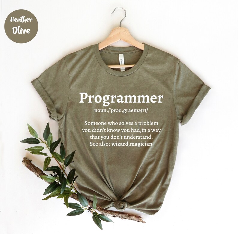 Puede incluir: Camiseta verde oliva con la palabra "Programmer" en blanco y su definici&oacute;n. La camiseta est&aacute; colgada en una percha de madera. Hay vegetaci&oacute;n en la parte inferior izquierda.