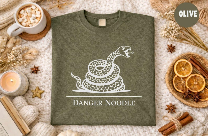 Peut inclure: T-shirt vert olive avec un motif de serpent blanc enroul&eacute; et l'inscription "DANGER NOODLE". Le t-shirt est pr&eacute;sent&eacute; sur une surface textur&eacute;e, avec des &eacute;l&eacute;ments d&eacute;coratifs.