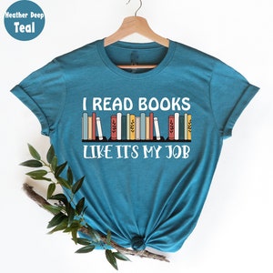 May include: Teal t-shirt with the text "I READ BOOKS LIKE IT'S MY JOB" in white. The design includes a row of colourful books. The shirt is displayed on a wooden hanger against a white background.