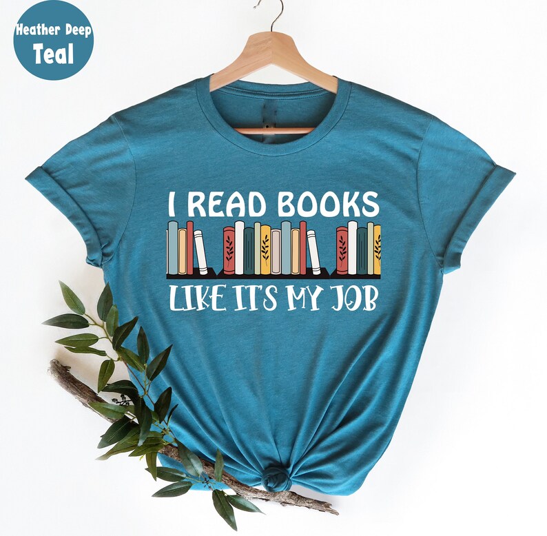 May include: Teal t-shirt with the text "I READ BOOKS LIKE IT'S MY JOB" in white. The design includes a row of colourful books. The shirt is displayed on a wooden hanger against a white background.