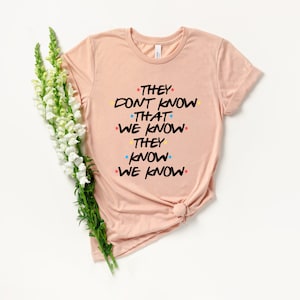 May include: A light pink t-shirt with the text "They don't know that we know they know we know" in black font with colorful dots.